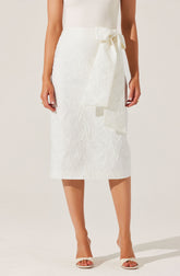 A woman is wearing a white, textured, midi-length skirt with a large side bow tied at the waist. She is also wearing white open-toe high heels.